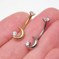Two crescent moon belly piercings, one in gold and one in silver, each featuring a clear cubic zirconia gemstone, shown resting on a person's fingers.