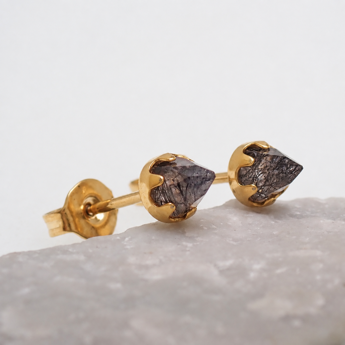 A pair of gold tourmalated quartz cone earrings resting against a stone with a white background 