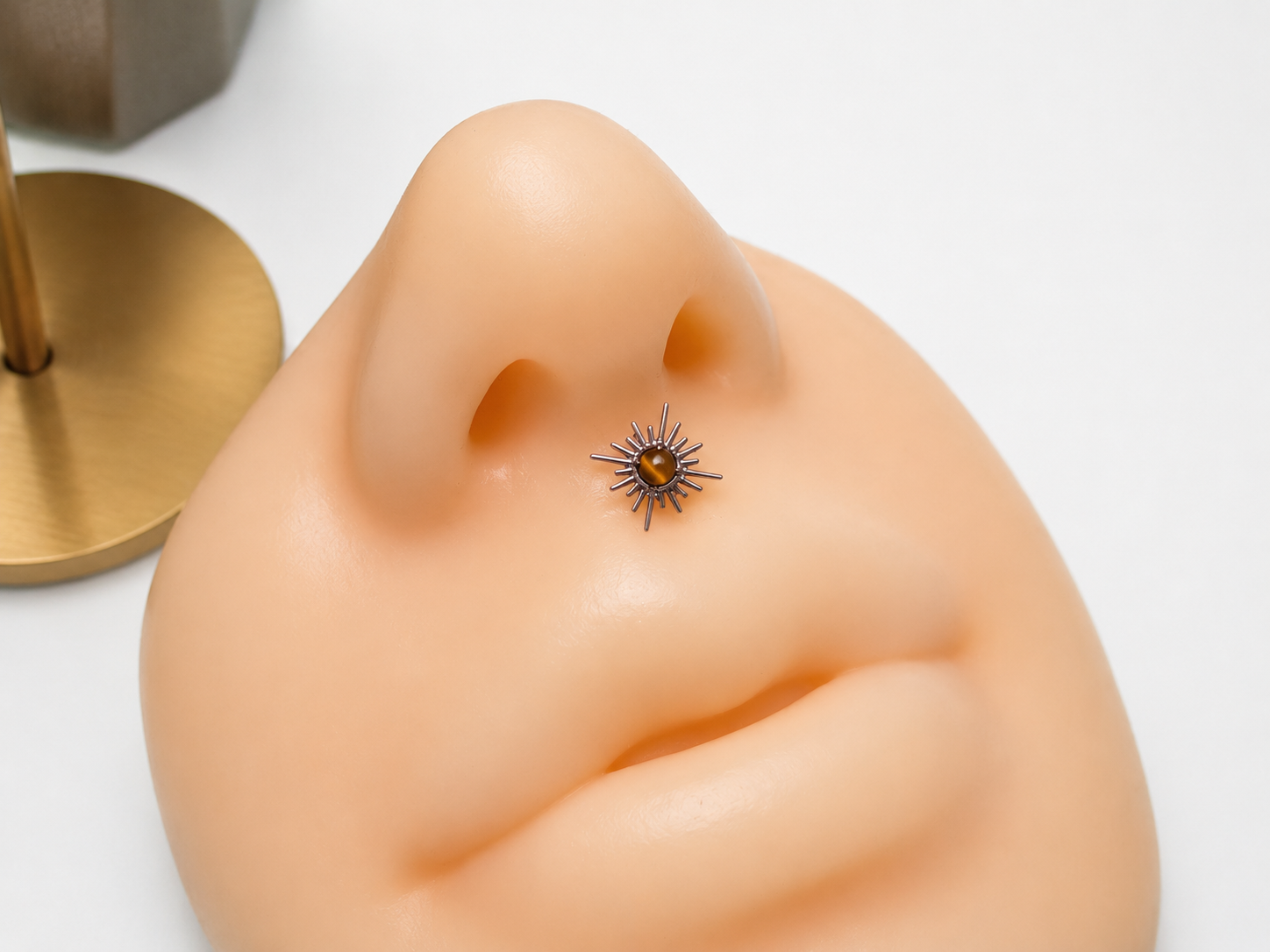 A close-up of a small metallic nose stud with multiple thin dark spikes and a central gold-toned oval stone, shown on a beige silicone nose model against a white background.