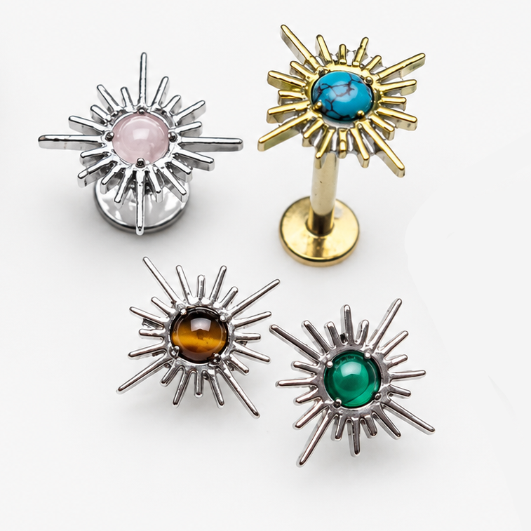 Four sunburst-style stud earrings arranged on a white background, with two gold-tone and two silver-tone earrings, each featuring a different colored gemstone (blue, green, amber, and pink). A green leaf and a rough stone are placed as styling props.