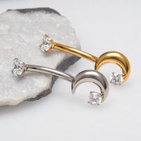 Two curved barbell nose studs with crescent moon ends, one in yellow gold and one in silver, each set with a clear cubic zirconia stone, displayed on a white surface with a polished stone prop.