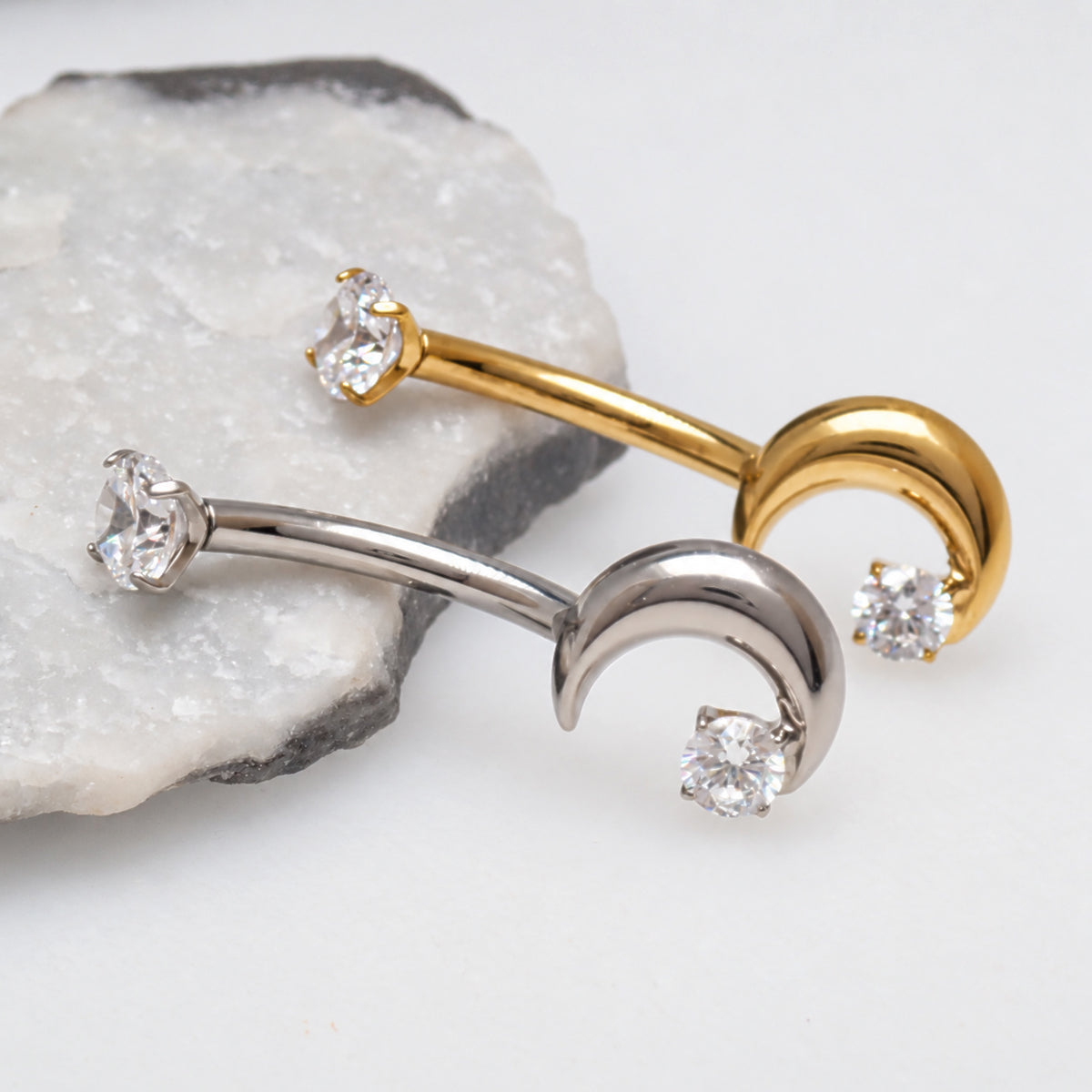 Two curved barbell nose studs with crescent moon ends, one in yellow gold and one in silver, each set with a clear cubic zirconia stone, displayed on a white surface with a polished stone prop.