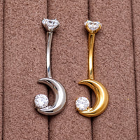 Two crescent moon belly button rings, one in silver and one in gold, both set with clear cubic zirconia stones, displayed on a brown textured background.