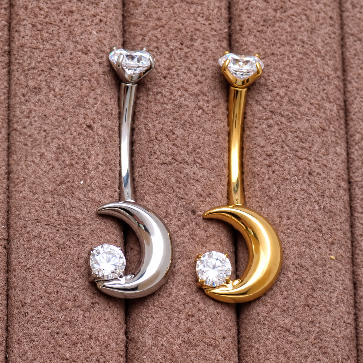 Two crescent moon belly button rings, one in silver and one in gold, both set with clear cubic zirconia stones, displayed on a brown textured background.