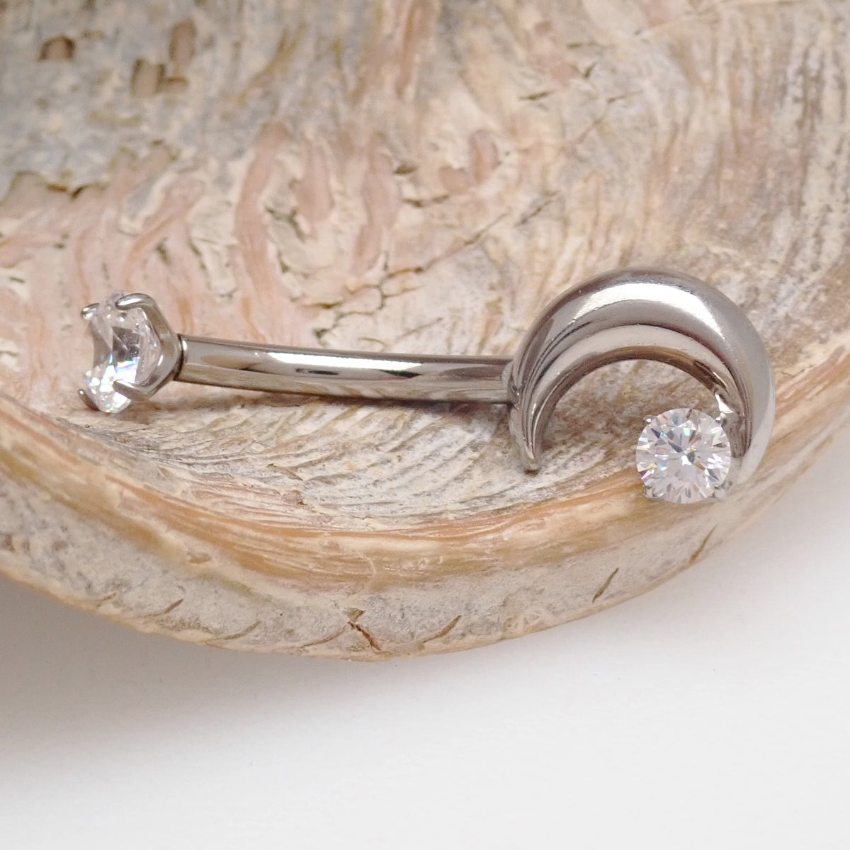 A silver crescent moon belly piercing on a wooden display with a white background 