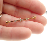 Gold industrial scaffold piercing with clear CZ and Trinity stones, featuring tri-flower design and titanium bar in 14g size.